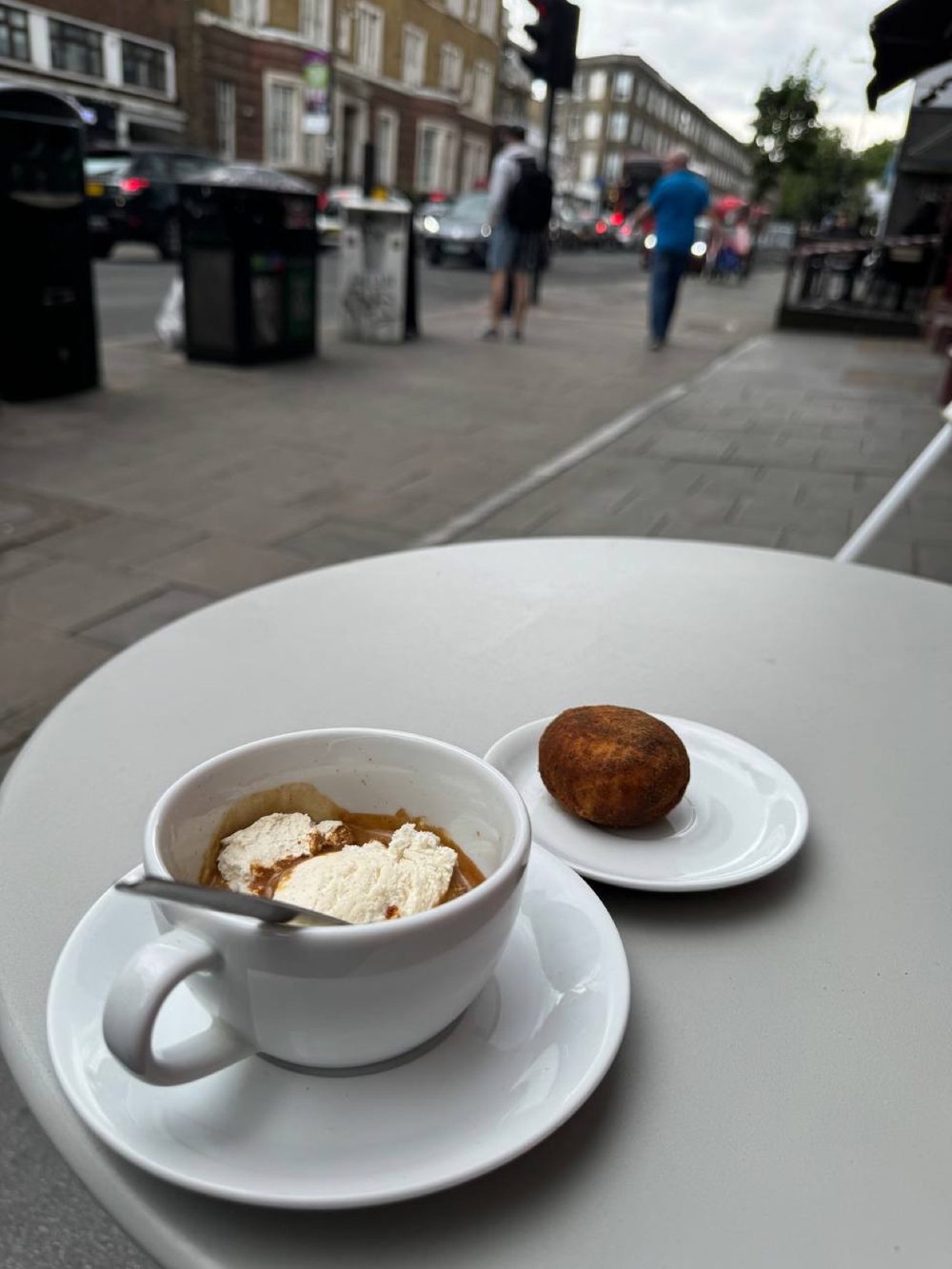 An outdoor pavement table with a coffee and a sugared bun, the busy street behind