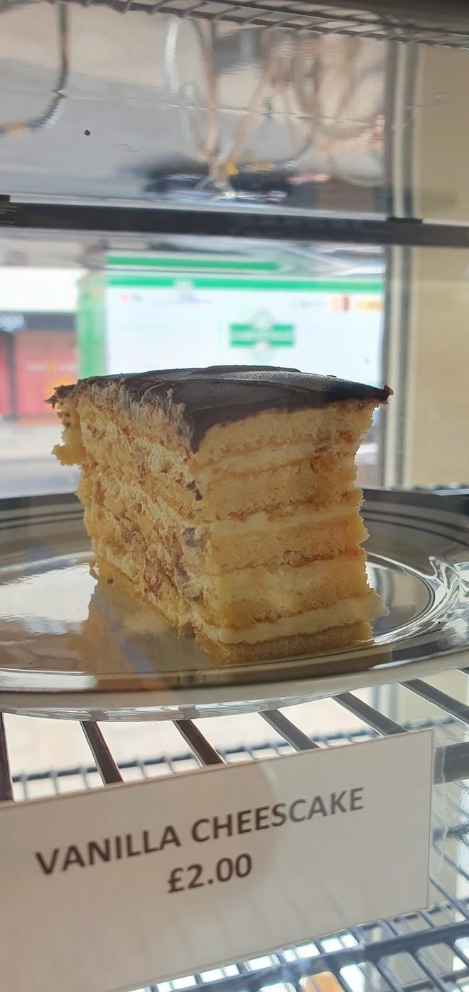 A layered chocolate-topped cake slice in the counter display case, cropped tight to the cake so the printed price card is out of frame